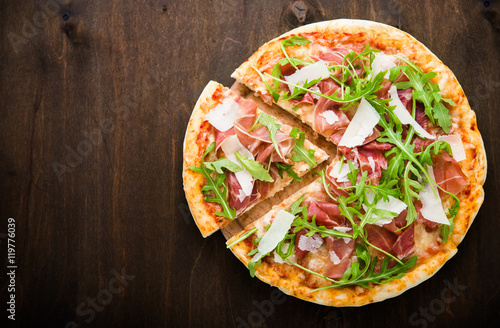 Foto Sliced pizza with prosciutto (parma ham), arugula (salad rocket) and parmesan on dark wooden background top view