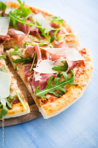 Sliced pizza with prosciutto (parma ham), arugula (salad rocket) and parmesan on blue wooden background close up. Italian cuisine.