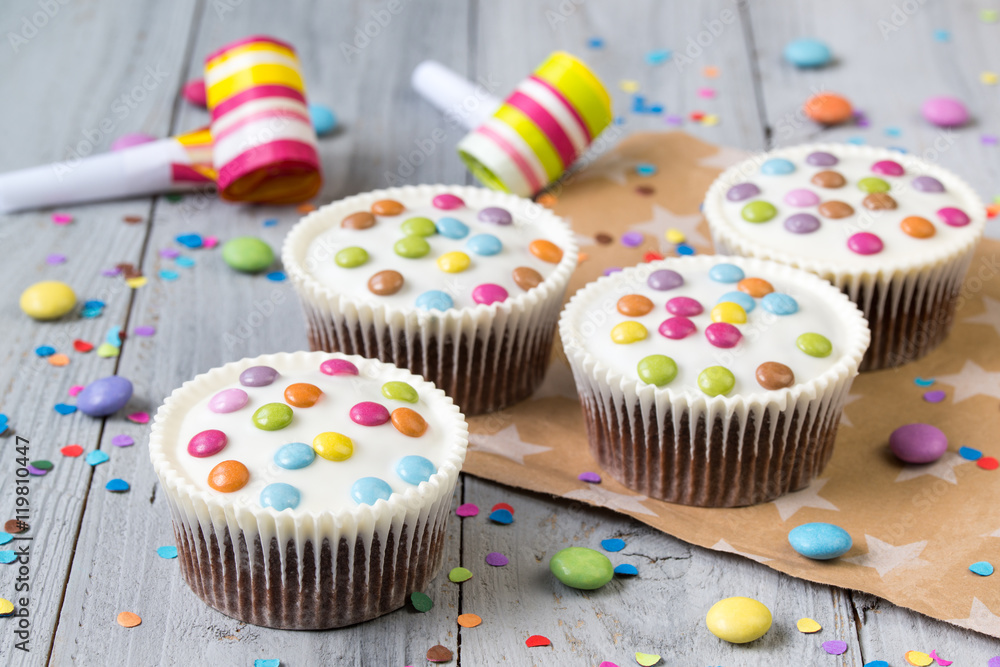 Cupcakes with white icing and colored smarties, party horns and confetti, wooden background Cupcakes with white icing and colored smarties, party horns and confetti, wooden background