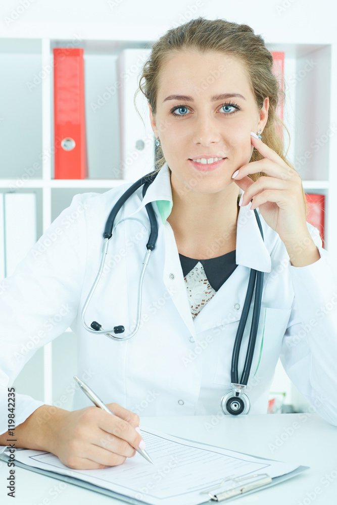 Beautiful young therapeutist sitting in front of working table, smiling and looking into camera. Medical concept.