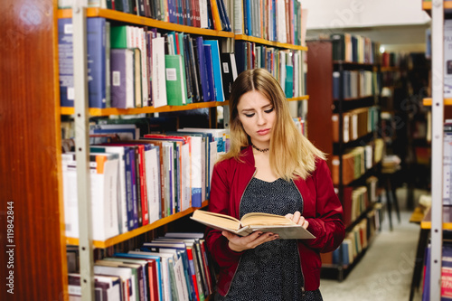 Female student reading a book between bookshelves in university library