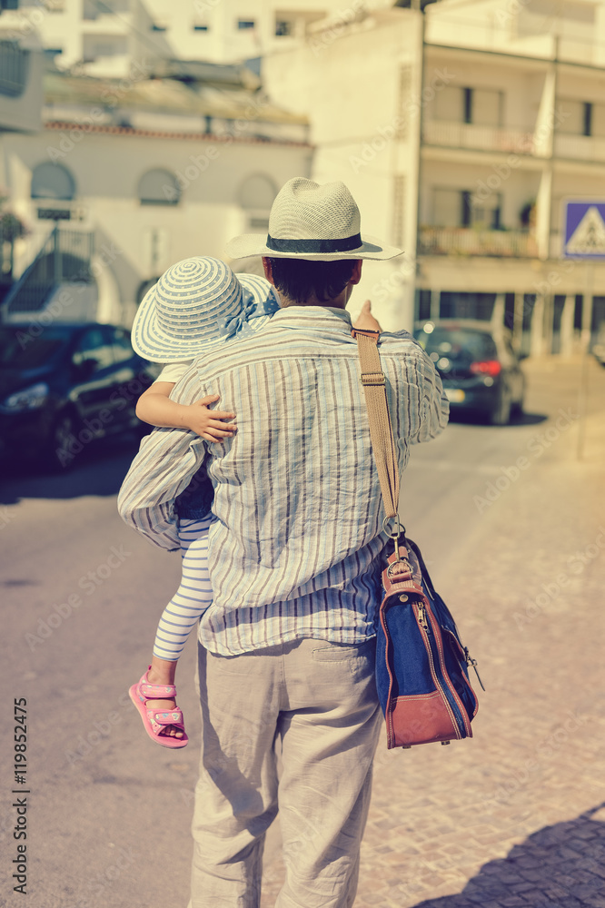Rear view of father holding child daughter walking the city street, sunny outdoors background