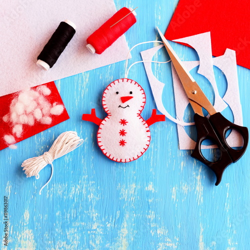 Cute felt Christmas snowman ornament, scissors, thread, needle, cord, felt sheets and scraps on wooden background. Supplies for making handmade snowman. Christmas kids crafts idea. Top view photo