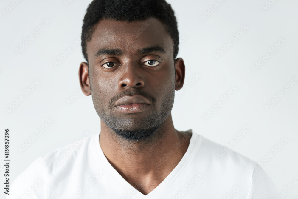 Obraz premium Highly-detailed close up portrait of good-looking young dark-skinned man wearing casual white T-shirt, looking at camera with serious and thoughtful expression on his face. Human emotions and feelings