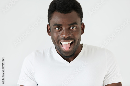 Human face expressions and emotions. Body language. Cheerful funny young African man wearing white T-shirt having fun, making faces, showing tongue to viewers, teasing them, spending free time indoors