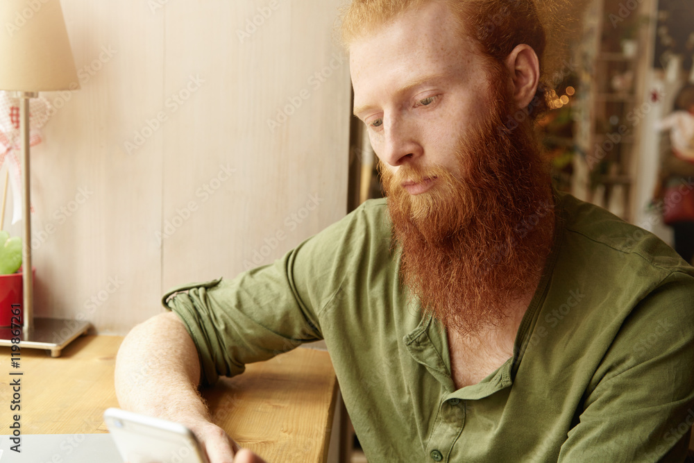 Obraz premium Headshot of young redhead male wearing green casual shirt, holding generic cell phone, browsing Internet and chatting with friends on electronic device, sitting against cafe interior background