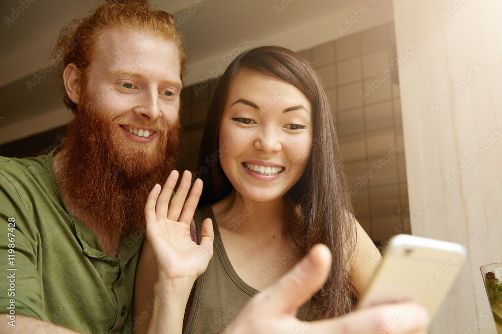 Young cute couple recording video for congratulating their friend on his birthday. Pretty student girl saying hello while taking selfie together with her redhead boyfriend using electronic device Young cute couple recording video for congratulating their friend on his birthday. Pretty student girl saying hello while taking selfie together with her redhead boyfriend using electronic device