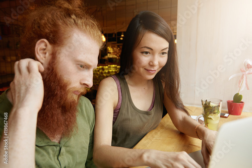 Two young colleagues working together on laptop computer, creating new business project: brunette girl with confident smile keyboarding, presenting her ideas to her companion with long red beard