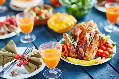 Rustic wooden table with abundance of food and dishes, focus on big golden roasted chicken and silverware decorated with berries and bows in autumn colors, pumpkin in background