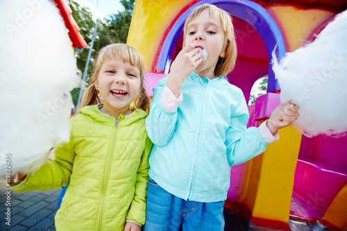 Portrait of two blond girls wearing colorful jackets on playground in amusement park holding big clouds of white cotton candy, eating it and smiling, looking at camera during family weekend