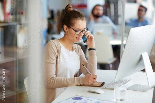 Young professional businesswoman working in public relations talking on phone with partners making notes in small notebook, sitting at computer desk in modern office space photo