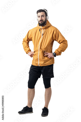 Jogger in sports jacket with earphones looking at camera in akimbo pose. Full body length portrait isolated on white studio background.