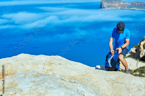 Fototapeta Naklejka Na Ścianę i Meble -  male traveler with backpack standing on the trail on the cliff against sea and blue sky at early morning. Balos beach on background, Crete, Greece