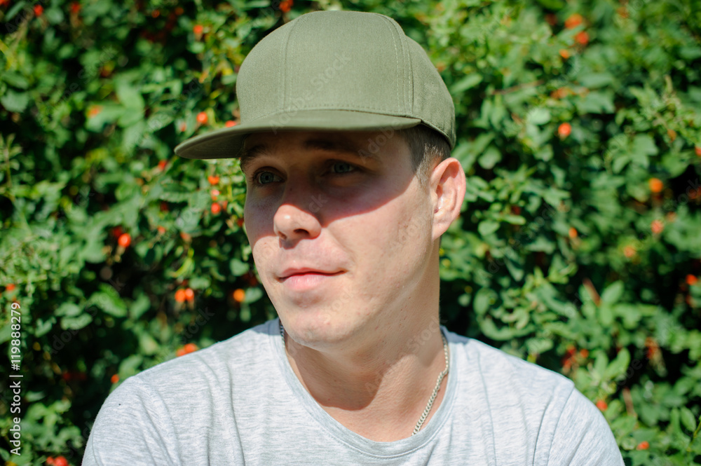 young guy standard physique posing on the street on a background of green bushes. He is wearing a light shirt and dark green cap. portrait of a man summer day