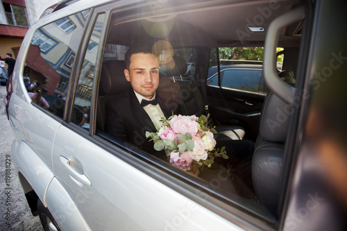 Handsome groom is sitting in car with bride's bouquet