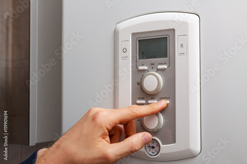 Men hand setting comfort temperature button on control panel of central heating or DHW at combi boiler in restroom.