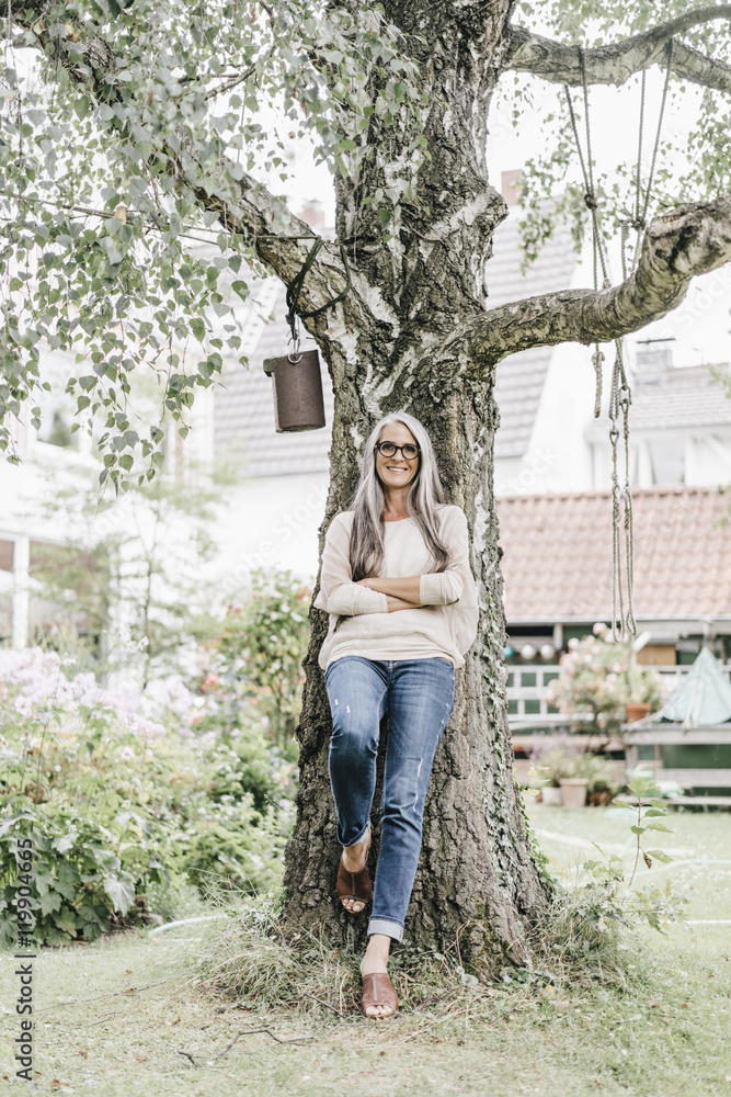 Smiling woman with long grey hair leaning against tree in the garden Smiling woman with long grey hair leaning against tree in the garden