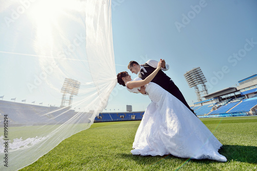 Wedding couple on the football stadium is hugging each other. Beautiful girl in white dress. Man in suit. Beauty bride with groom. Female and male portrait. Woman with lace veil. Lady and guy outdoors