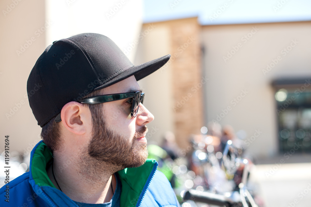 Portrait of young handsome man with black beard wearing dark sunglasses, cap and blue sleeveless jacket looking at the street on outdoor in the summer day Portrait of young handsome man with black beard wearing dark sunglasses, cap and blue sleeveless jacket looking at the street on outdoor in the summer day