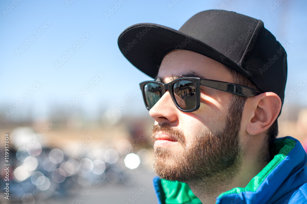 Portrait of young handsome man with black beard wearing dark sunglasses, cap and blue sleeveless jacket looking at the street on outdoor in the summer day Portrait of young handsome man with black beard wearing dark sunglasses, cap and blue sleeveless jacket looking at the street on outdoor in the summer day