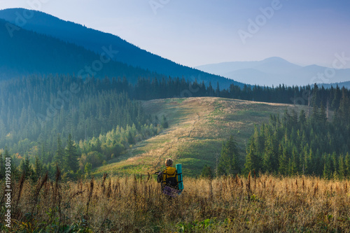 Backpacker is walking through tall grass in the autumn mountains. View on the wooded hills and hazy peaks in the distance.