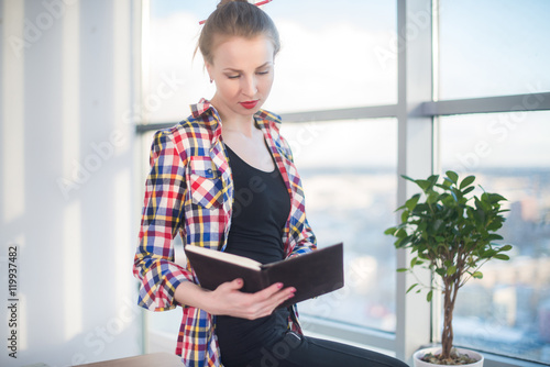 Side view portrait of young woman sitting, looking down, reading the book, learning at light room in morning with her torso turned.