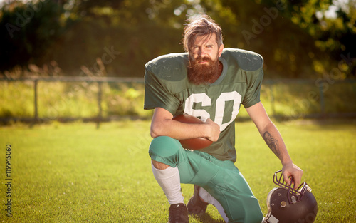 Brutal man with a long beard and mustache in the shape of an American football player with helmet and Ball on the training ground