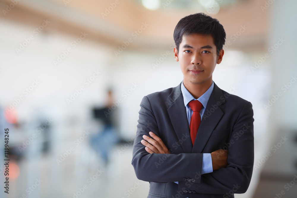 Cheerful young asian businessman in full suit keeping arms crossed and looking at camera in office Cheerful young asian businessman in full suit keeping arms crossed and looking at camera in office