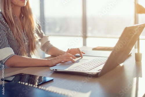 Woman blogging in spacious office using computer on her workplace. Female employee sitting, smiling, looking at camera.