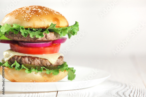 Big juicy traditional American burger with grilled meat and slices fresh vegetables closeup on a white plate on a wooden table. Food and beverages. Selective focus with shallow depth of field