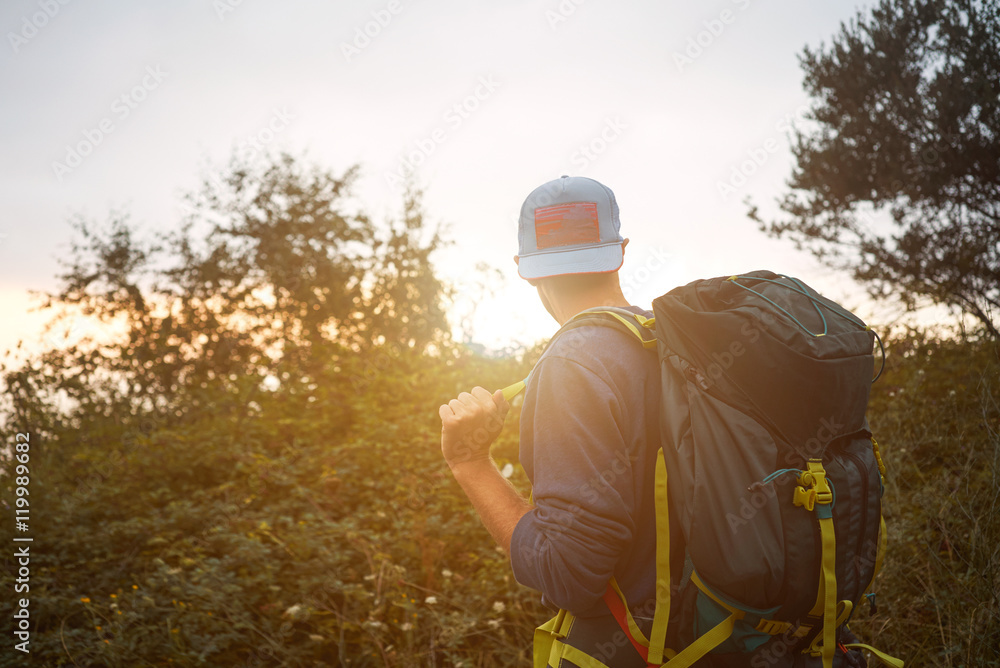 Hip young trekker walking on a hill among trees looking at sunset and carrying his big backpack Hip young trekker walking on a hill among trees looking at sunset and carrying his big backpack