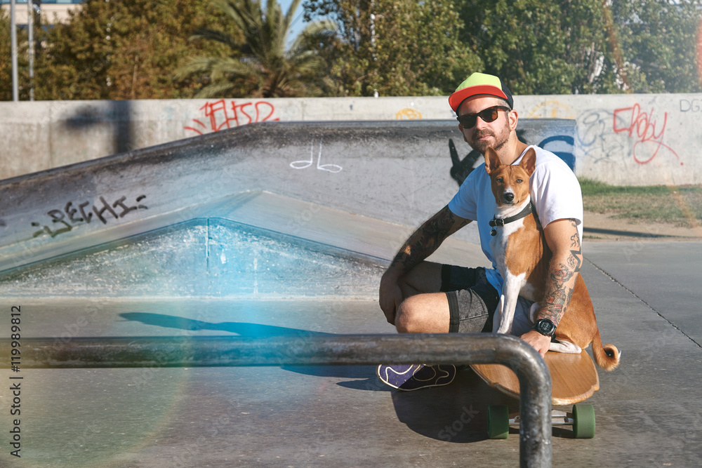 Φωτογραφία Surfer in baseball hat sits on his wooden longboard skate hugging his basenji dog in empty morning skate park pool and looking at camera Light leak styling