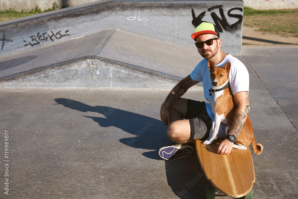 Φωτογραφία Surfer in baseball hat sits on his wooden longboard skate hugging his basenji do