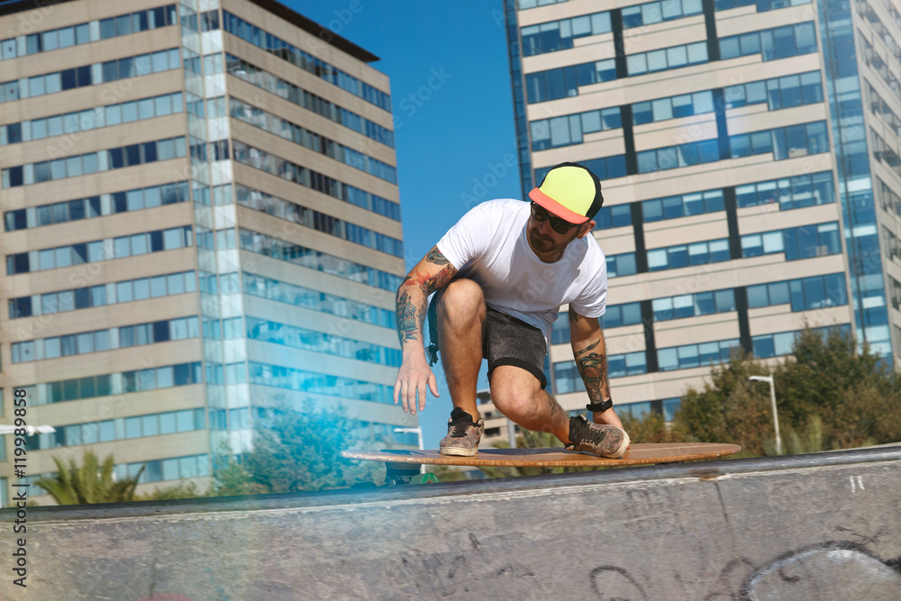 Φωτογραφία Tattooed skater in plain blank t-shirt makes short angle turn on his londoard on top of concrete pool in skatepark in front of urban landscape