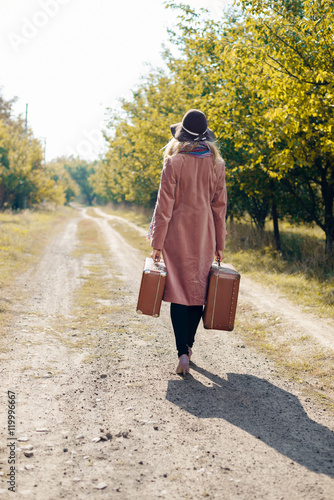 Back view of female wearing hat coat walking away on rural road. She carrying old suitcase over sunny background outdoors
