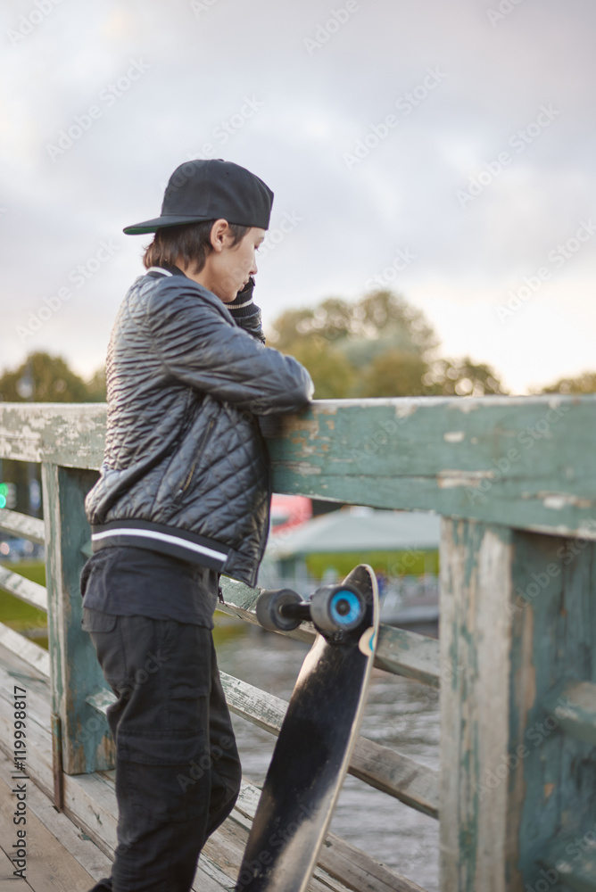 Girl or boy in dark wear stay on the wooden bridge and thinking or talk by phone after longboard or skateboard riding. Dreaming concept.