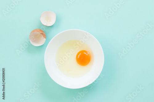 Top view of raw egg in white bowl with eggshell on blue background.