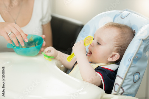 Cheerful little boy sitting in highchair and playing with spoon