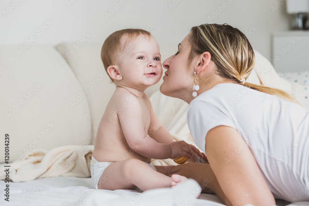 Beautiful young mother kissing baby boy in cheek on bed Beautiful young mother kissing baby boy in cheek on bed