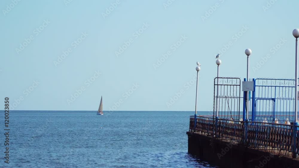 Luminaires are on the breakwater on the lamps sitting gulls. On the horizon sailing yacht at sea Luminaires are on the breakwater on the lamps sitting gulls. On the horizon sailing yacht at sea