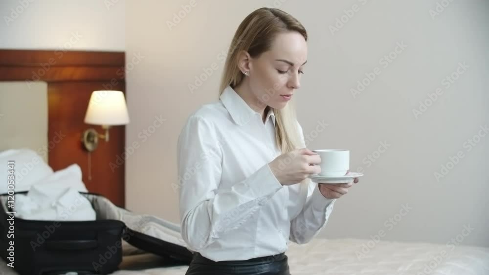 Medium shot of young businesswoman sitting on hotel bed and drinking coffee before morning meeting, unpacked suitcase lying in background Medium shot of young businesswoman sitting on hotel bed and drinking coffee before morning meeting, unpacked suitcase lying in background
