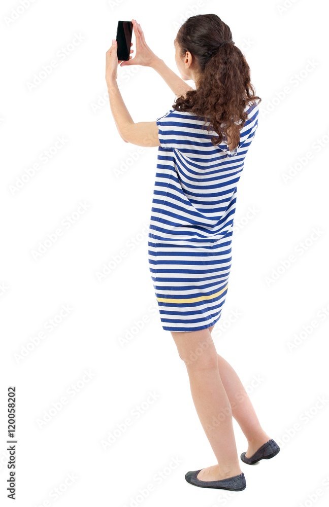 back view of standing young beautiful woman using a mobile phone. girl watching. Swarthy girl in a checkered dress takes on the phone. back view of standing young beautiful woman using a mobile phone. girl watching. Swarthy girl in a checkered dress takes on the phone.