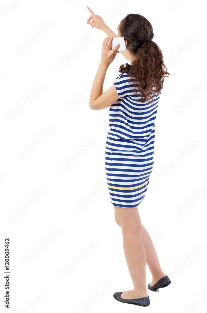 back view of standing young beautiful woman using a mobile phone. Swarthy girl in a checkered dress shows a finger and talking on the phone. back view of standing young beautiful woman using a mobile phone. Swarthy girl in a checkered dress shows a finger and talking on the phone.