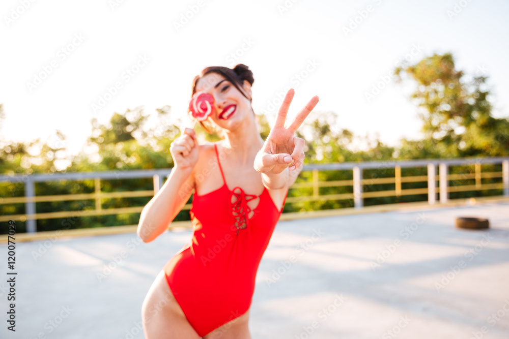 Happy woman covered eye with lollipop and showing peace sign