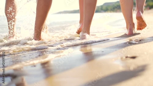 Legs of group of friend while barefoot walking on coast. Sea waves, foams and sprays. Tracking slow motion shot