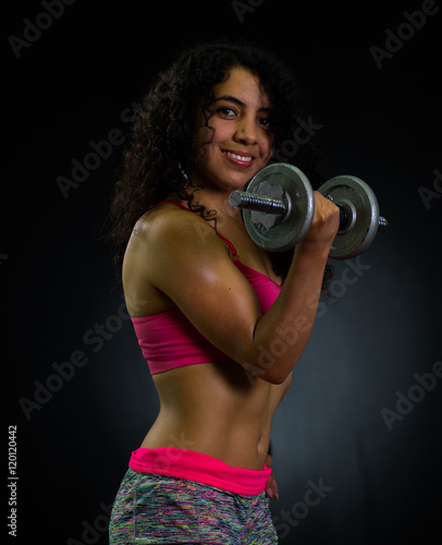 Athletic brunette woman wearing pink top and matching shorts, training with weights while posing for camera, black studio background