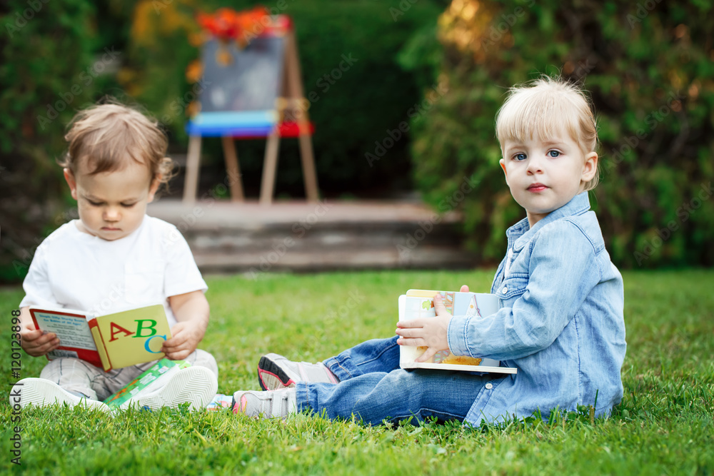 Group of two white Caucasian toddler children kids boy and girl sitting outside in grass in summer autumn park by drawing easel with books, playing studying learning, back to school Group of two white Caucasian toddler children kids boy and girl sitting outside in grass in summer autumn park by drawing easel with books, playing studying learning, back to school