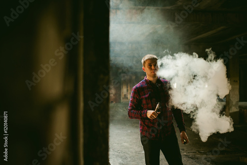 man in a plaid shirt and jeans, smokes an electronic cigarette, blowing smoke and steam from the vaporization a mechanical device. location Abandoned unfinished building, brick walls, stylish.