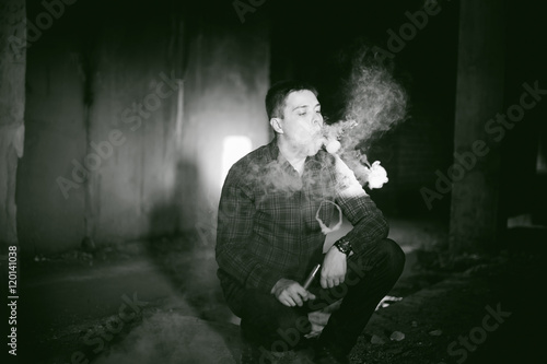 man in a plaid shirt and jeans  smokes an electronic cigarette  blowing smoke and steam from the vaporization a mechanical device. fashionable hobby  alternative to smoking cigarettes  glycerin clouds