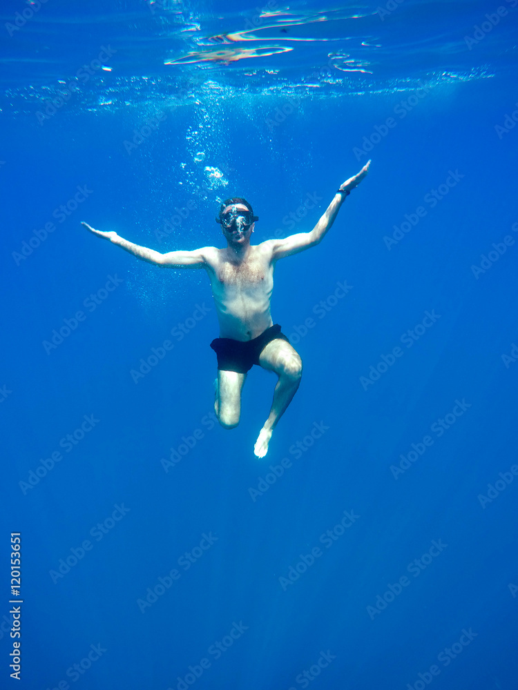 Portrait of a young causasian man meditating in the lotus position underwater. Submerged under water free diving, crossed legs, wearing a mask and blowing bubbles.